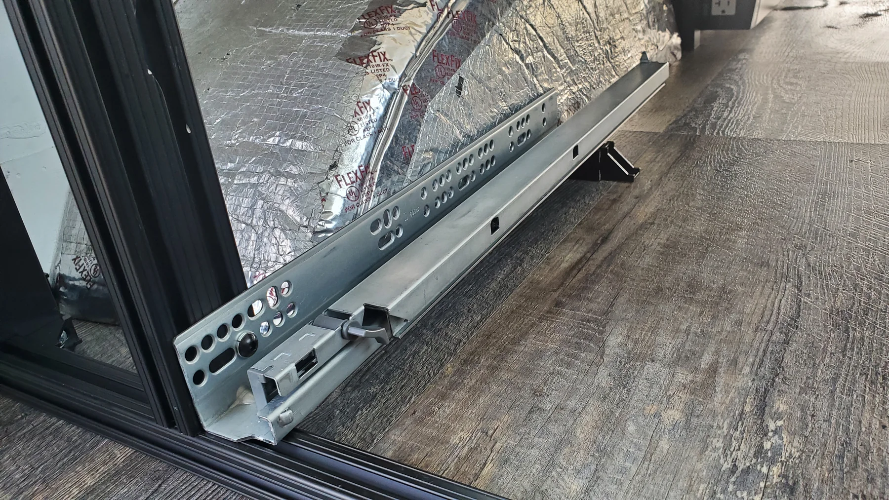 Metal drawer runner mounted low inside a black aluminum frame opening, beside an insulated wheel well.