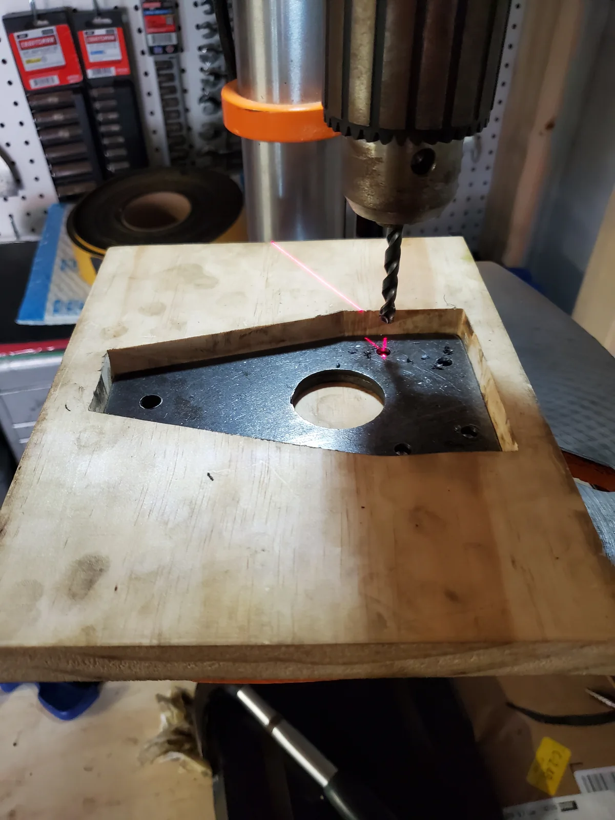 Dark metal plate in a routed wooden pocket beneath a drill press, with holes and metal chips visible.