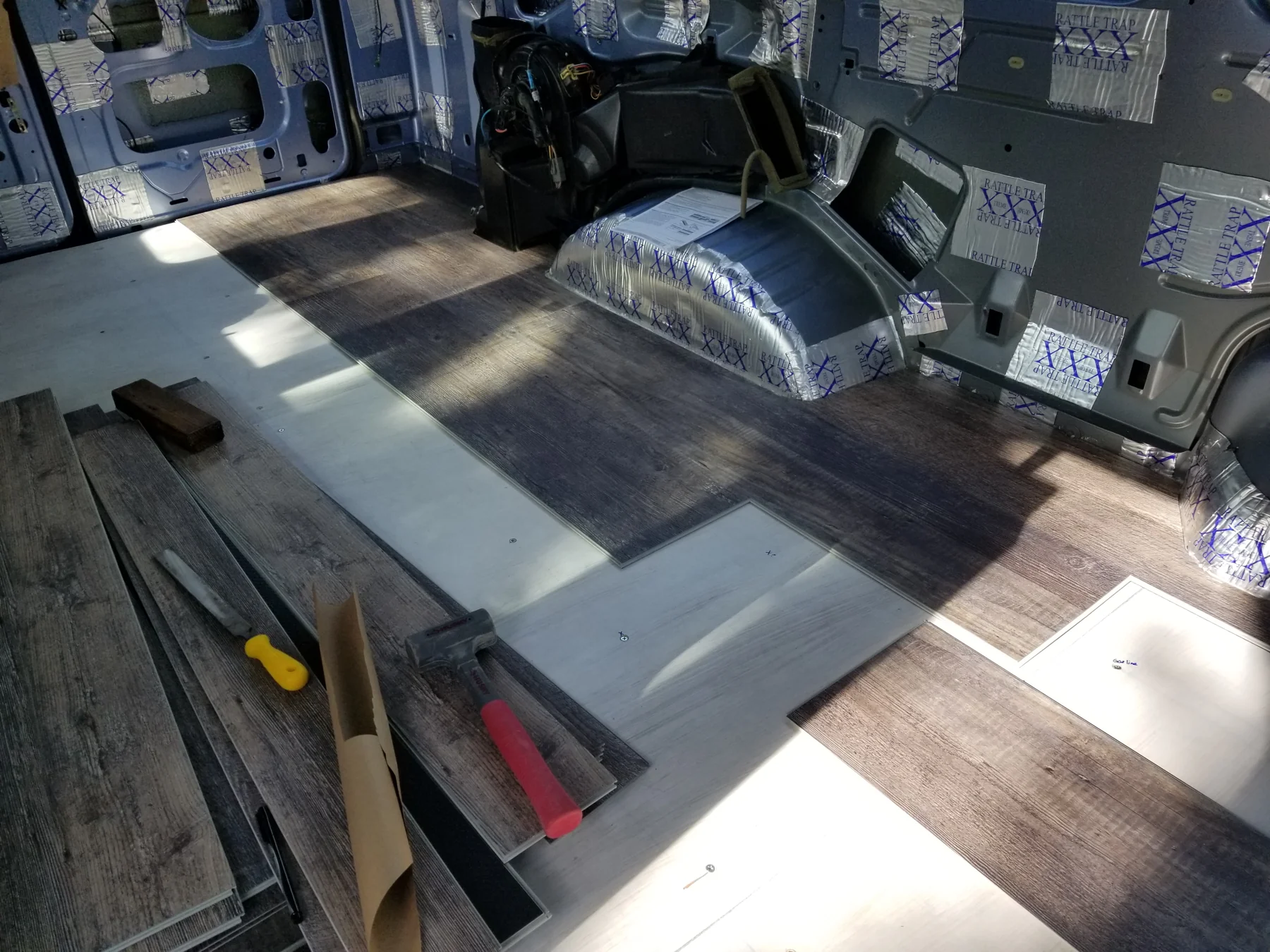 Staggered wood-pattern flooring boards being fitted around a wheel well, with tools and spare boards nearby.