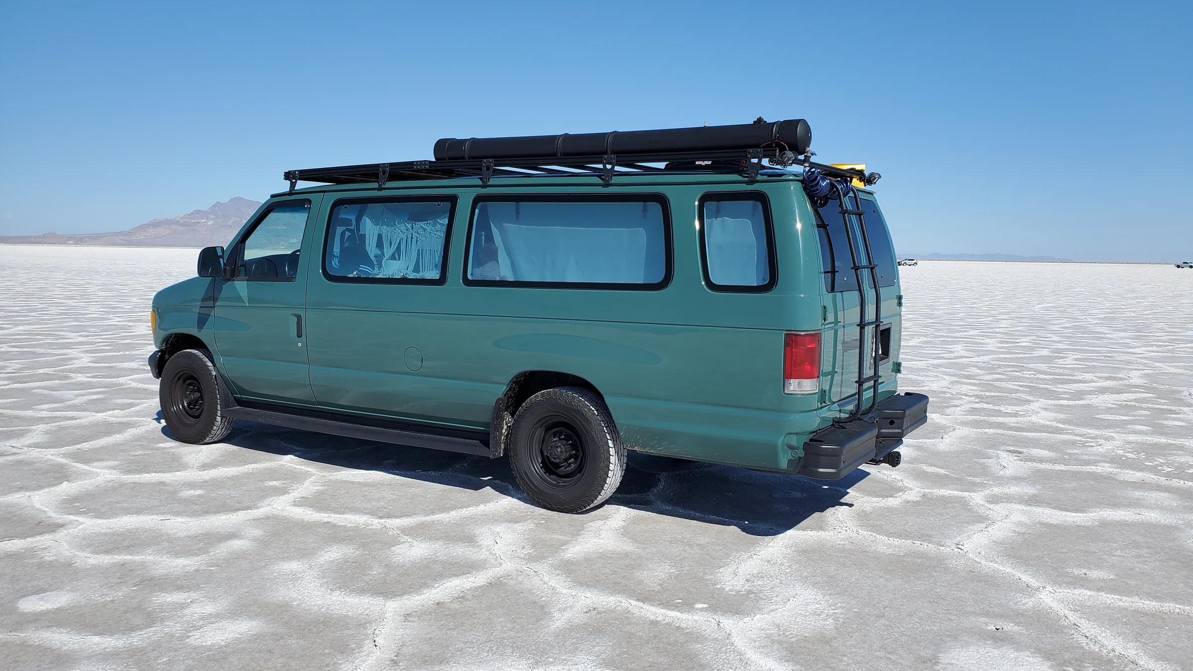 Green Ford Econoline campervan with a roof rack on an open white landscape under a blue sky.