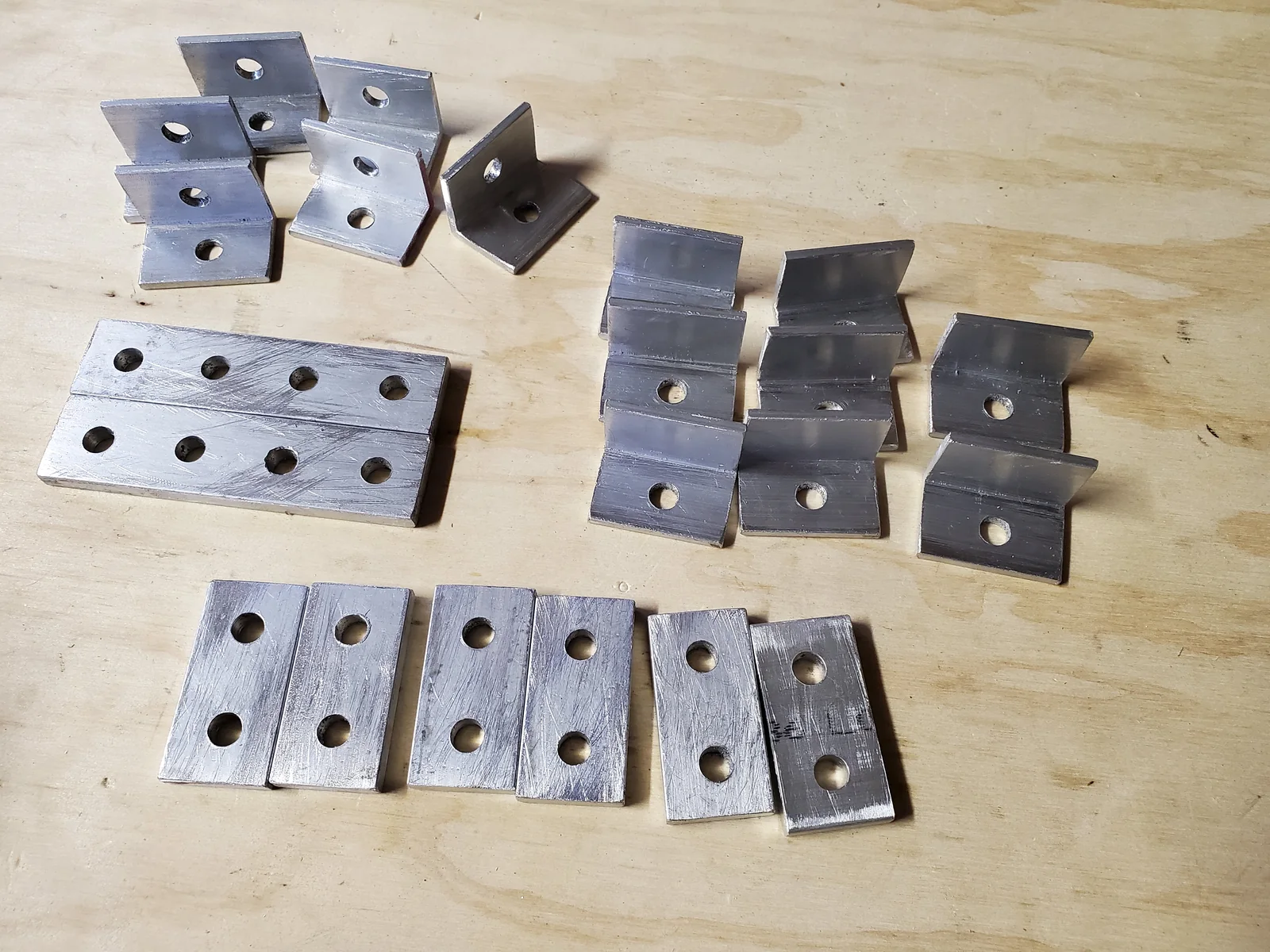 Rows of bent metal brackets and flat drilled plates arranged on a wood surface.