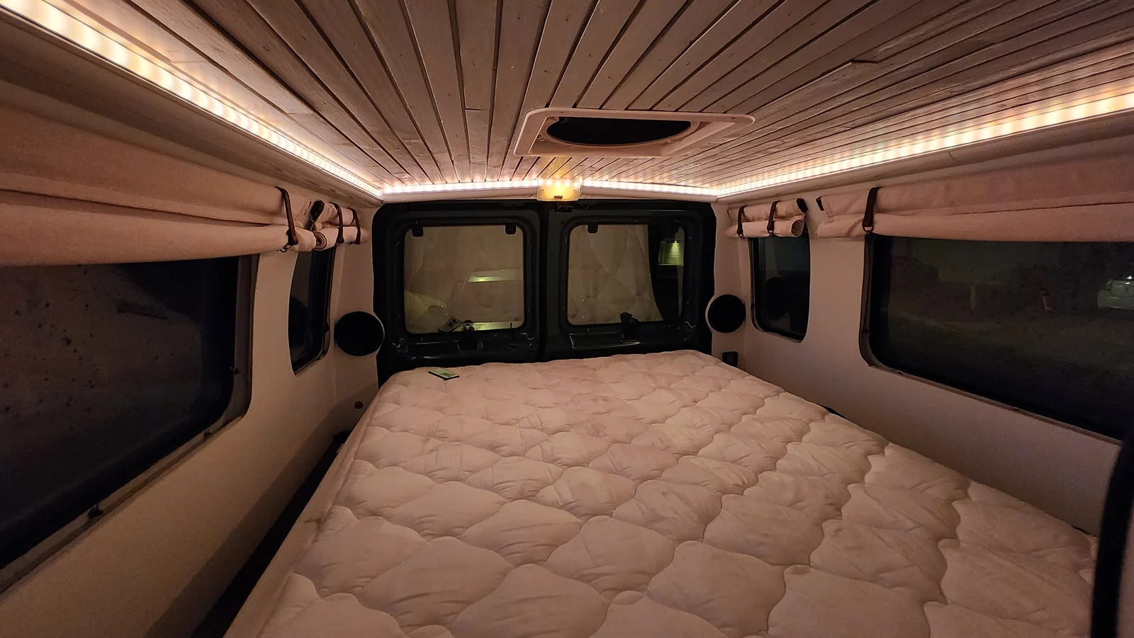 Campervan bed and wood ceiling illuminated by warm LED lights at night.
