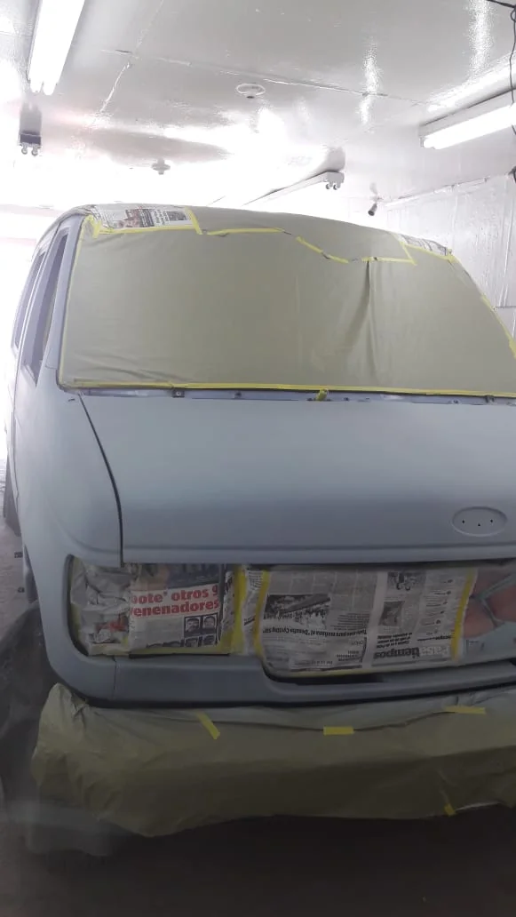 Gray van front inside a booth with windshield and front openings masked by paper and tape.
