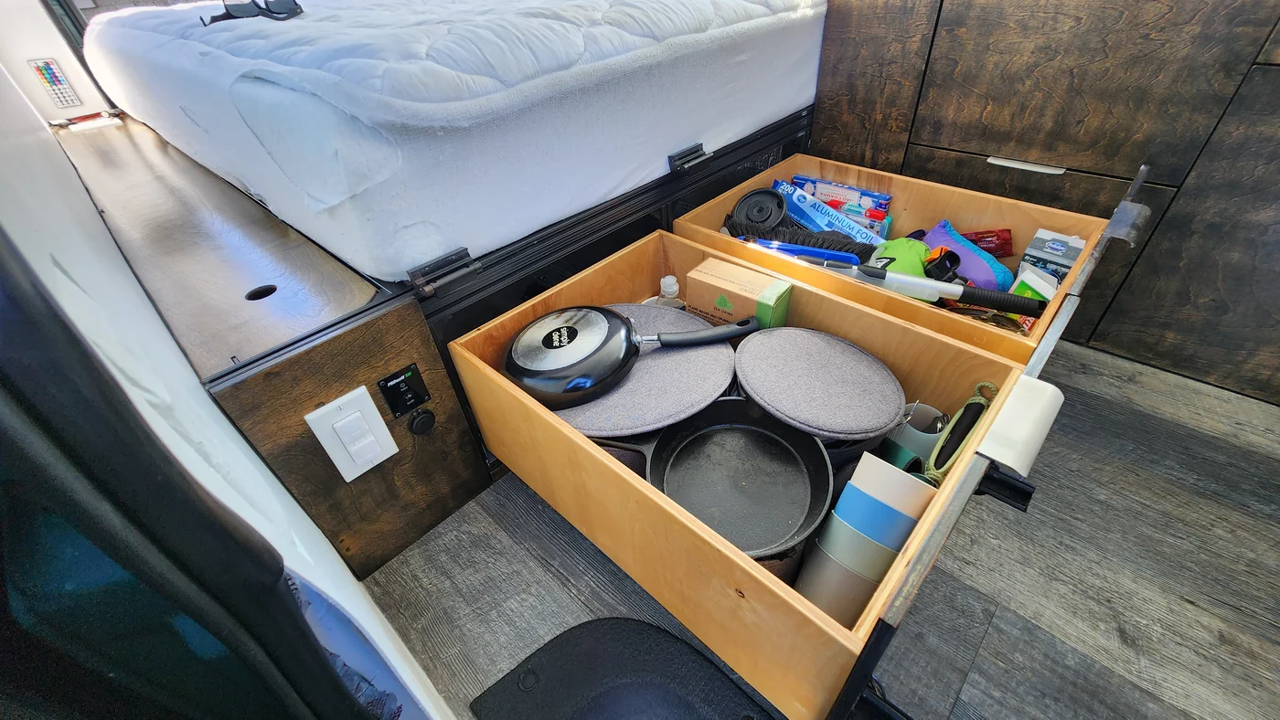 Wide under-bed drawer pulled out into the campervan aisle with cookware inside.